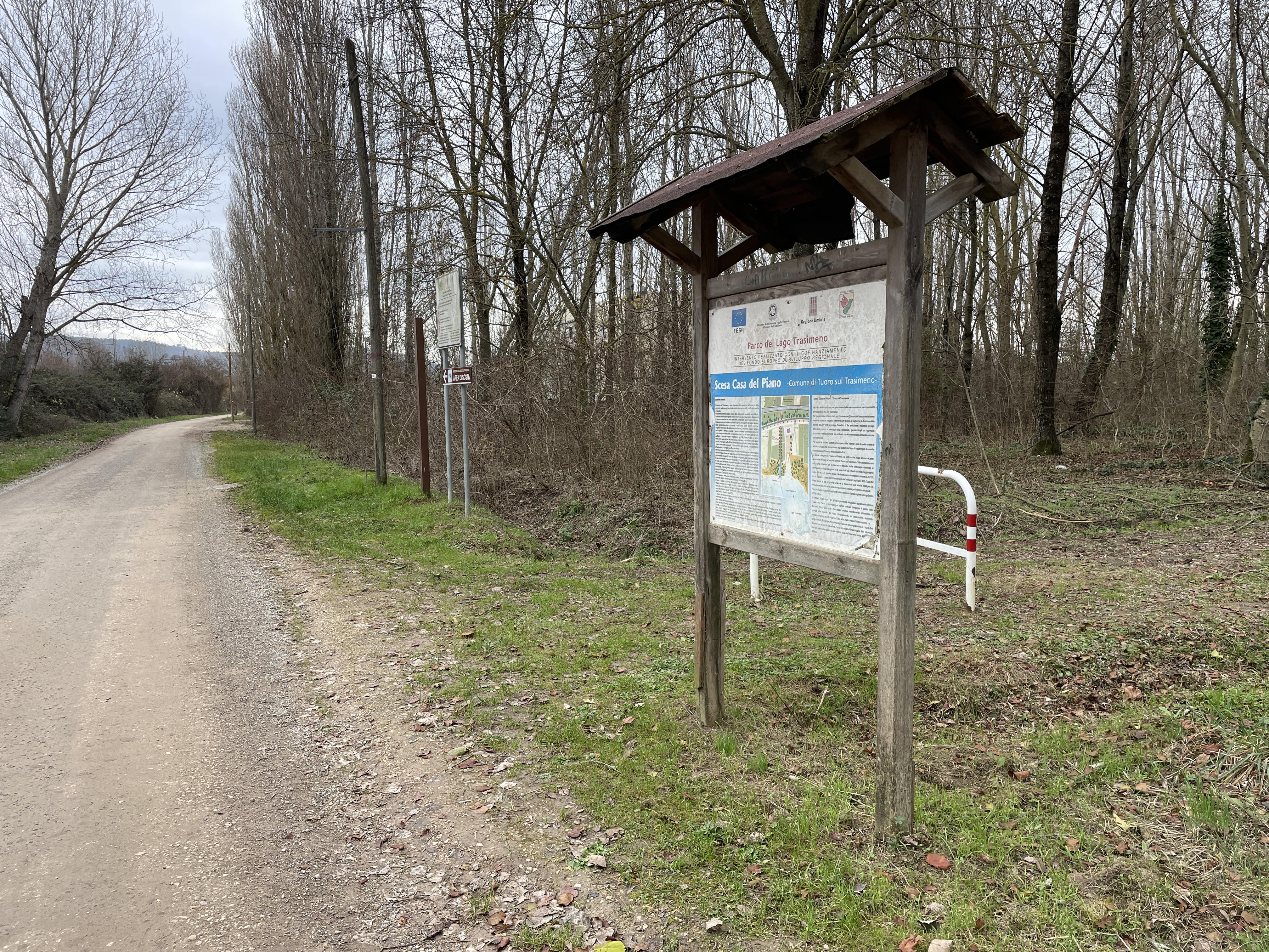 Radweg auf unbefestigtem Weg mit kahlen Bäumen. Rechts ein Informationsschild über den Park und die Oase Casa del Piano.